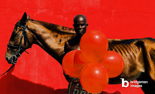 Photo of a man standing in front of a horse, red background Derrik Boateng / Bridgeman Images